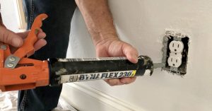 A picture of a man using caulk to seal the gaps around an electrical outlet with the cover off. This helps for energy efficiency by air sealing your home.