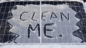 A picture of two solar panels together with light snow covering them. Written in the snow is "clean me", indicating the need to brush off the snow.