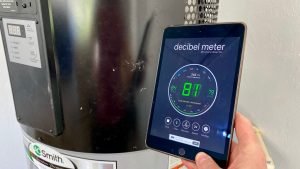 A picture of our hybrid heat pump hot water heater in the background, with me holding up an iPad. The screen shows a decibel level reading of 81, indicating how loud this hot water heater is.