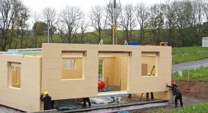A picture of a small prefabricated home being built. It shows unpainted walls hanging from a crane being moved into place with the help of two construction workers.