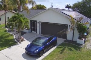 A picture of a blue Tesla Model 3 in front of our white-painted net-zero house with solar panels on the roof, and palm trees around the yard.