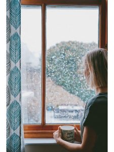 Photo of a woman in shirt sleeves holding a mug of tea looking out the window of a house at snow falling