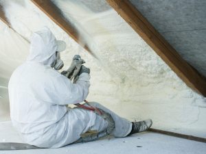A man in full protective gear is sitting on the attic floor while he sprays spray foam insulation on the underside of the attic ceiling.