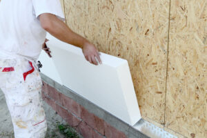 A picture of a person in a white construction outfit installing rigid foam board insulation on a wood wall.