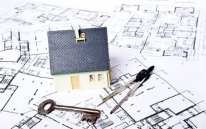 house planning and architectural drawings on a table, with a small model house, key, and protractor laying on top of them