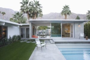 a small white modern house with a pool in the foreground, and California mountains in the background with palm trees