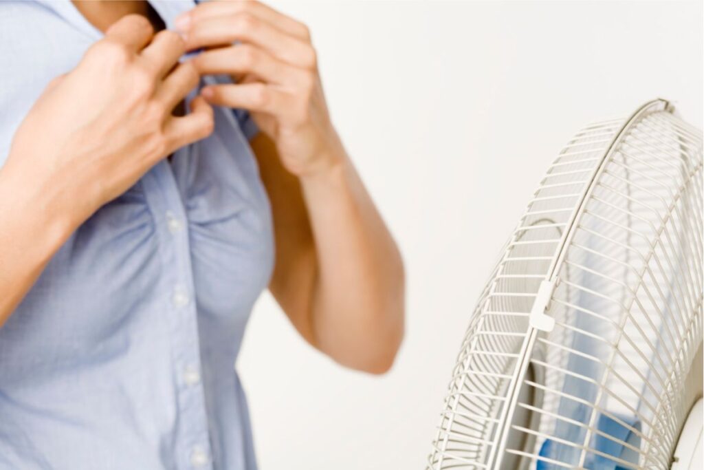 A woman standing in front of an oscillating fan unbuttons her blouse to cope with high humidity levels