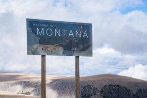 The stateline road sign in the foreground welcomes visitors to Montana with mountains visible in back