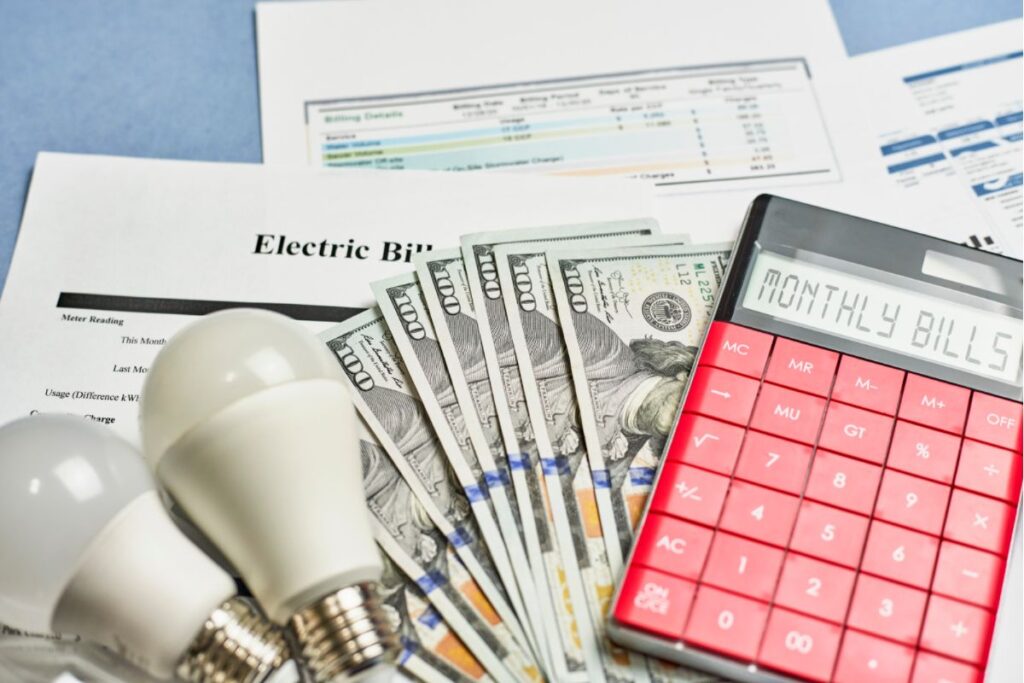 A calculator and light bulbs stacked on top of a bundle of cash and utility bills on a table