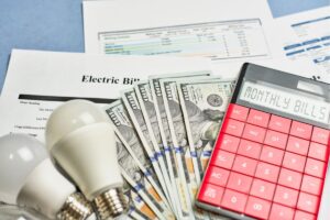 A calculator and light bulbs stacked on top of a bundle of cash and utility bills on a table