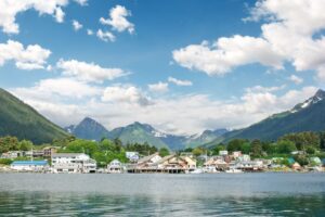 A town in Alaska with buildings along the waterfront and mountain peaks and cloudy, blue skies in back