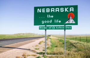 The Nebraska state sign at the state line along a highway with fields and blue sky in the background