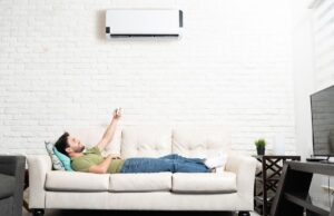 A homeowner laying on his living room couch uses a remote to adjust the setting of the mini-split air handler on the wall above
