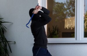 A burglar facing an exterior hurricane window of a home winds up to smash it with a crowbar