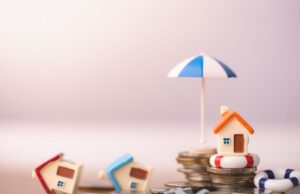 Two tipped-over model homes, and one upright on a stack of coins, protected by an umbrella, indicating insurance savings from protective measures