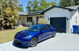 a blue tesla model 3 parked on the grey driveway with a white solar netzero home