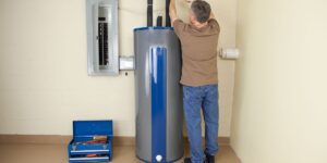 Blue and gray hot water cylinder in the corner of a room showing water and electrical connections. Technician is working on the inlet pipe with his back to the camera. There is a toolbox on the ground to the left.