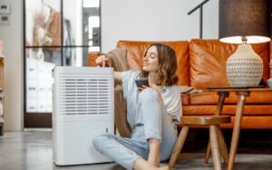 A homeowner sits on her living room floor in front of the couch with her arm resting on a dehumidifier