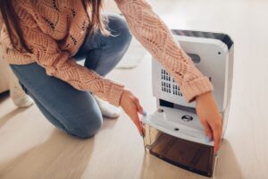 Woman crouching next to a portable dehumidifier to empty the condensate bucket. This is to illustrate maintenance that can influence how long do dehumidifiers last.