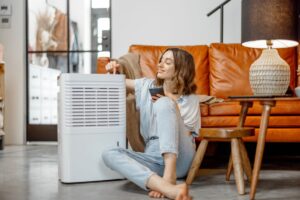 A homeowner sitting in front of her living room couch smiles as she rests one arm on a dehumidifier