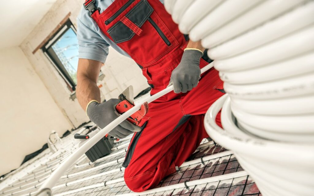 A technician kneeling on the floor cuts a piece of cable from a spool during a radiant floor heating system installation