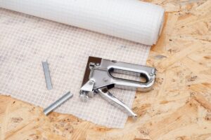 A vapor barrier roll, staple gun, and some staples resting atop a piece of plywood