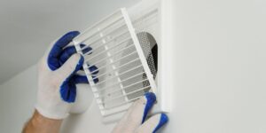A white vent set into a white painted wall with a person wearing blue and white gloves replacing the screen.
