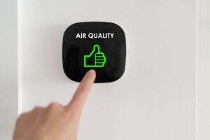 A black button mounted on a white wall with the words, "Air Quality" written in white text above a green "thumbs up" icon. The button is being pressed by a person's index finger.