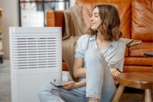 A homeowner sitting on the floor in front of her living room couch peers down at the dehumidifier to her right