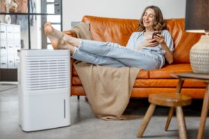 A homeowner sitting on her couch, smiling as she looks at and rests her feet on a dehumidifier