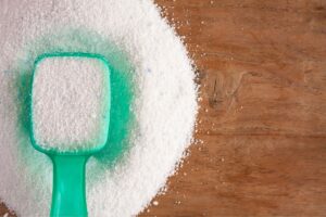 A pile of laundry detergent on a wooden tabletop and a semi-full scoop on top of the pile