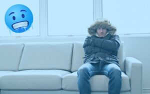 a man sitting on a couch inside wearing a big winter coat and looking unhappy