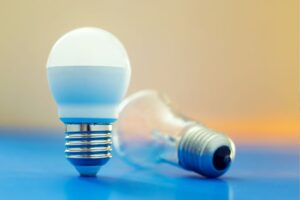 A incandescent e26 bulb leaning against an LED light bulb on a blue tabletop.