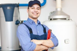 A smiling technician with arms crossed standing between a conventional and a heat pump water heater