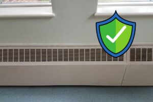 The vent of a baseboard heating system running along a wall with a green safety check graphic at the top right corner.