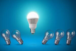 An illuminated LED bulb against a blue background towering above a row of incandescent bulbs