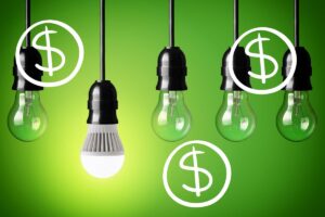 An illuminated LED bulb hanging lowest in a row of all-incandescent bulbs, with multiple dollar signs superimposed above the image