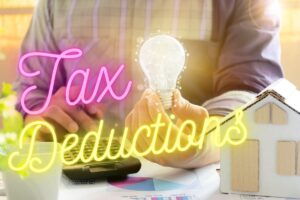 A homeowner sitting at a desk holding an illuminated energy-saving light bulb in one hand and punching numbers into a calculator with the other, with the text, "tax deductions" superimposed above the image.